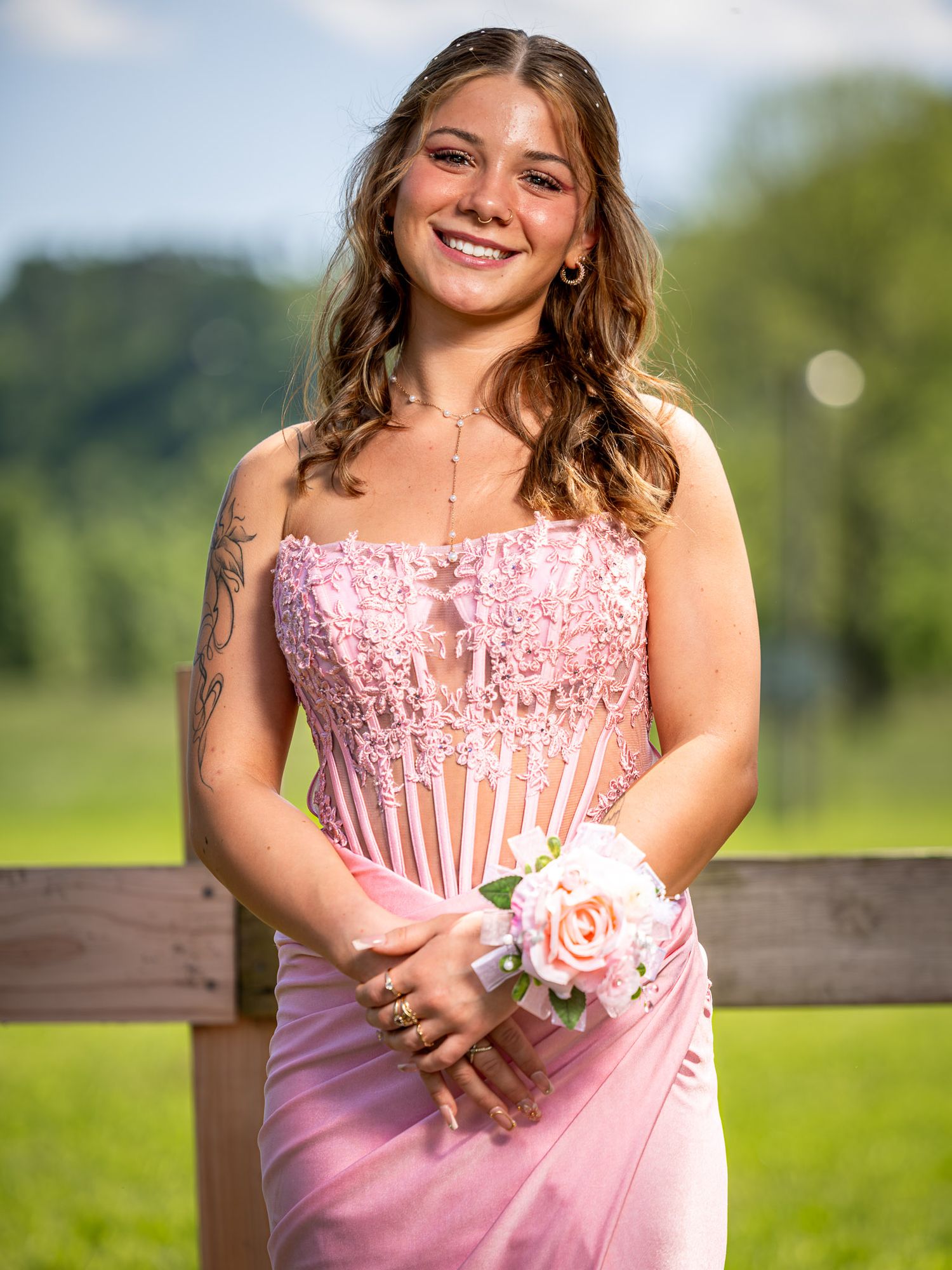 Young woman in pink lace corset prom dress holding floral wrist corsage, smiling outdoors near wooden fence.