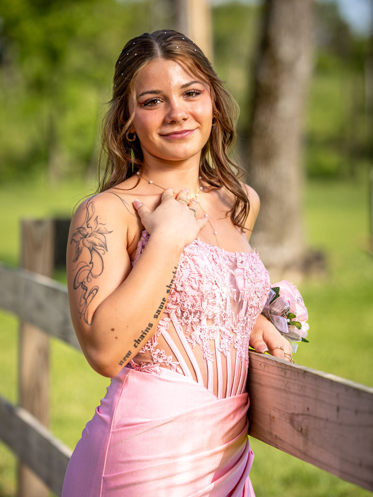 Young woman in pink lace prom dress with floral tattoo holds corsage by wooden fence outdoors.