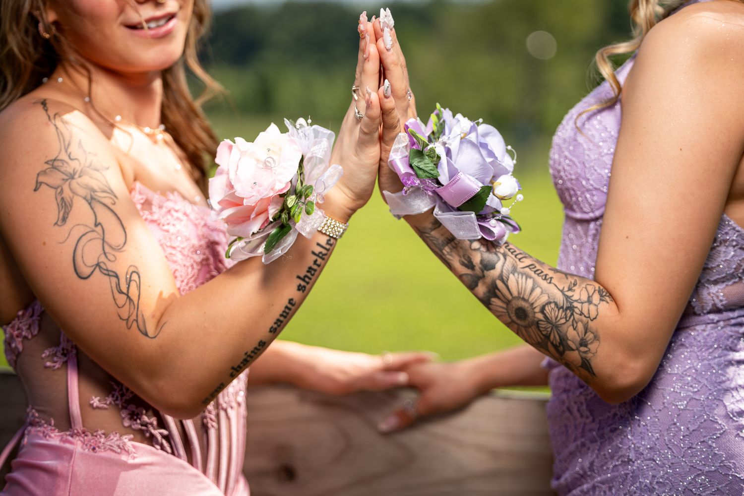 Two tattooed women in pink and purple formal dresses holding floral corsages together outdoors.