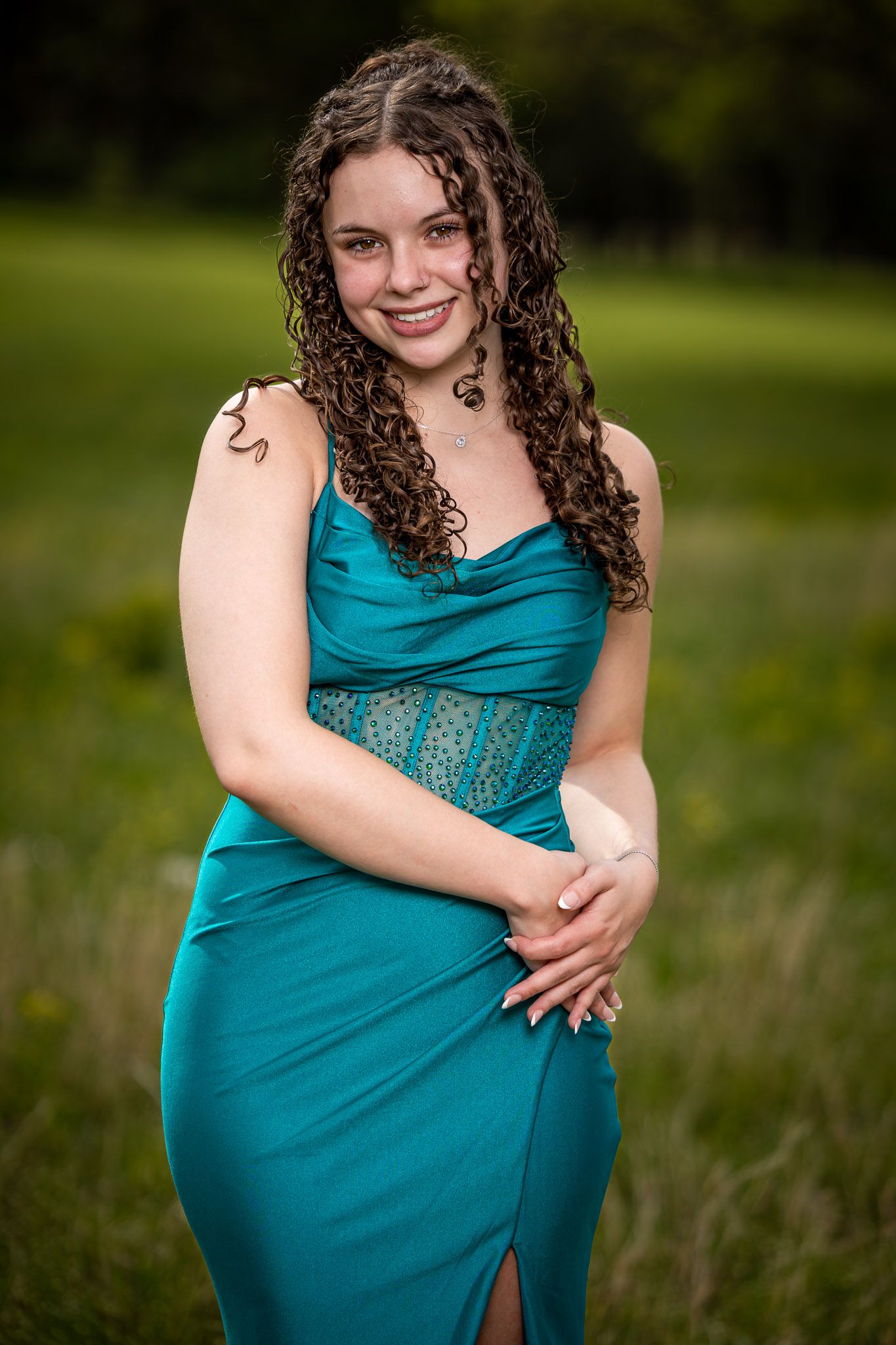 A smiling young woman in a teal satin gown with lace detail poses outdoors in a green field.