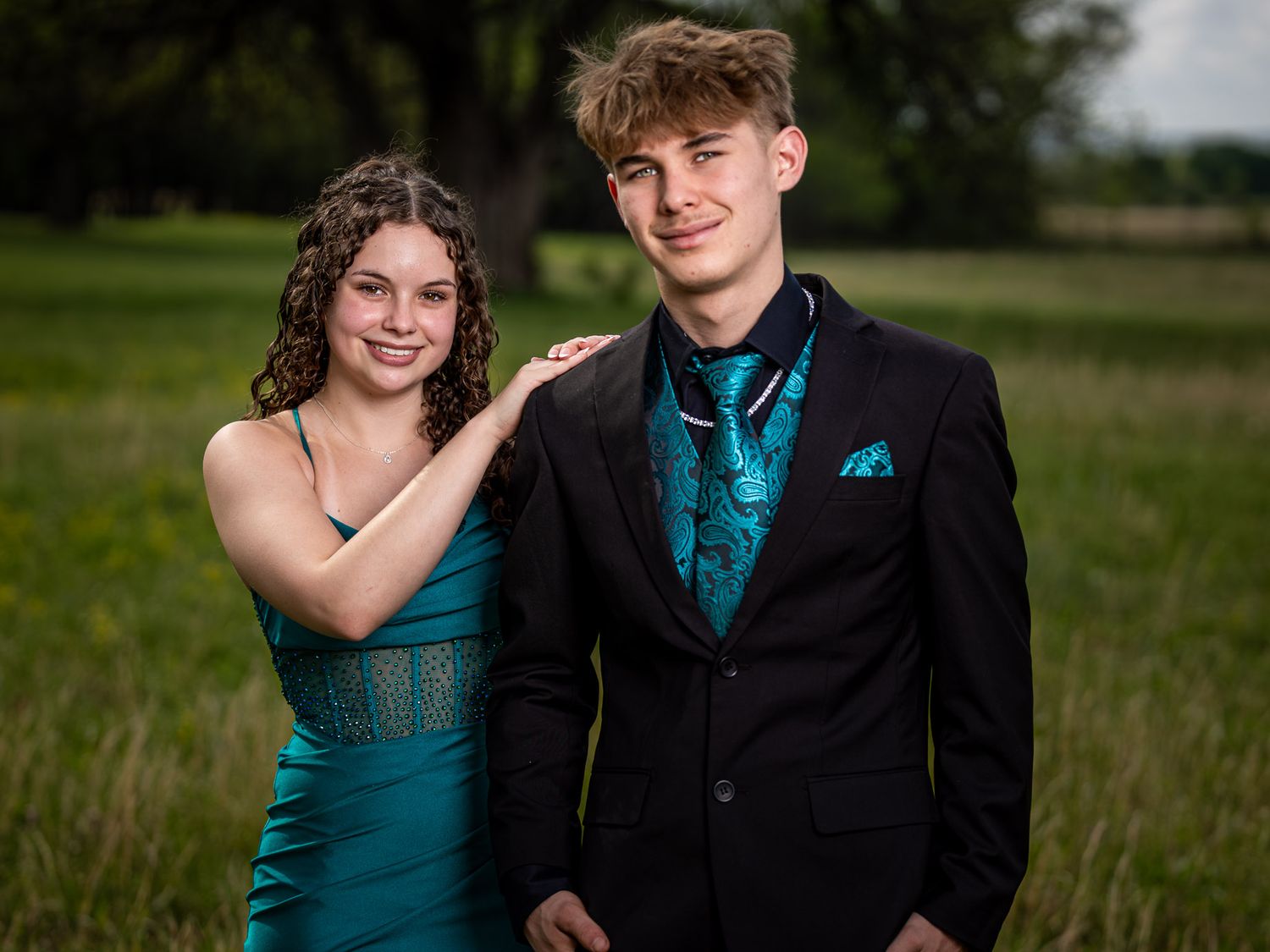 A teenage couple dressed in matching teal formal wear poses outdoors in a green field for prom photos.