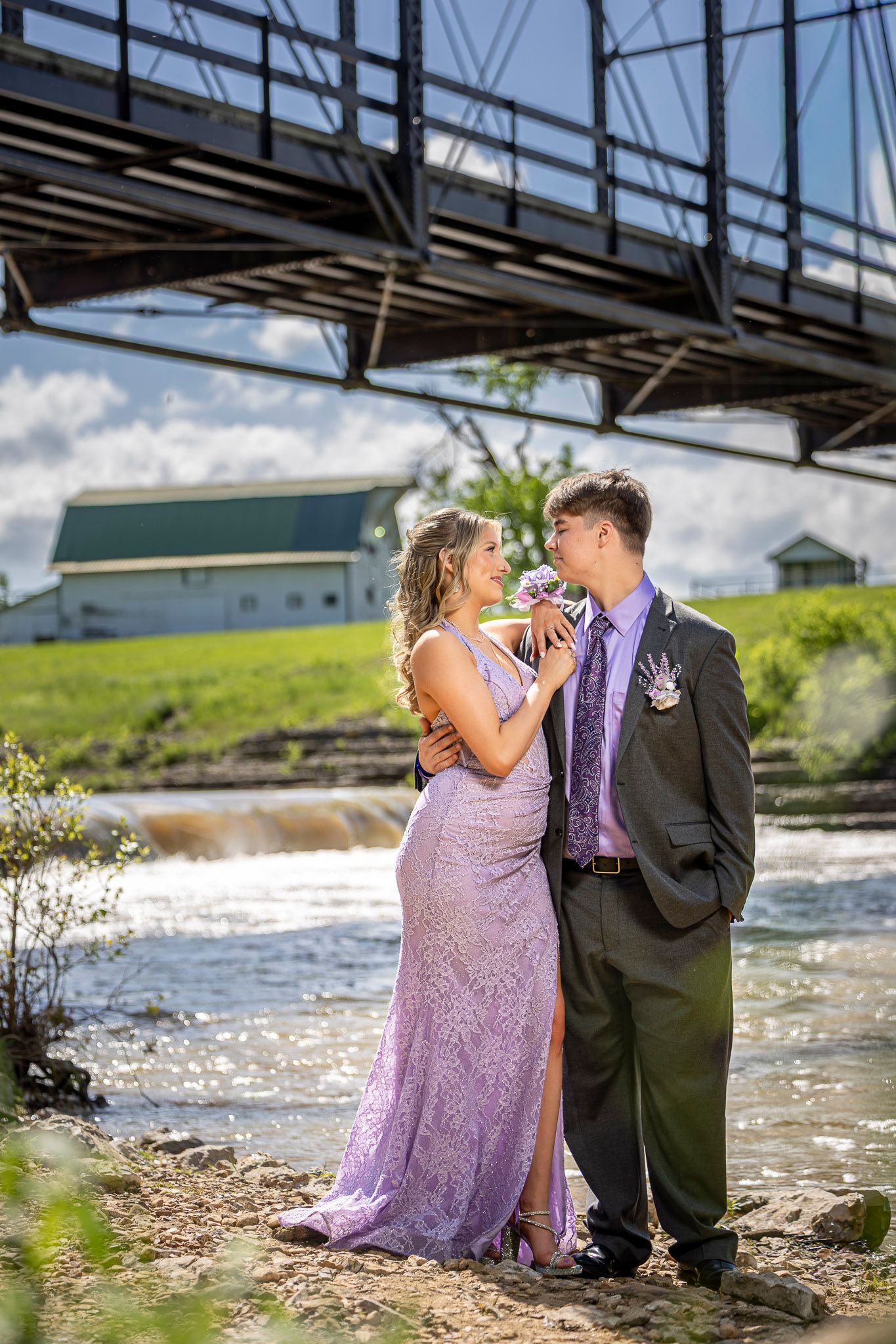 A couple in prom attire poses by a river under a bridge, she in lavender gown, he in dark suit with purple tie.