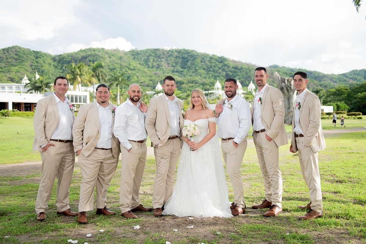Bride and groom pose with seven groomsmen in tan suits on tropical resort lawn with mountain backdrop.