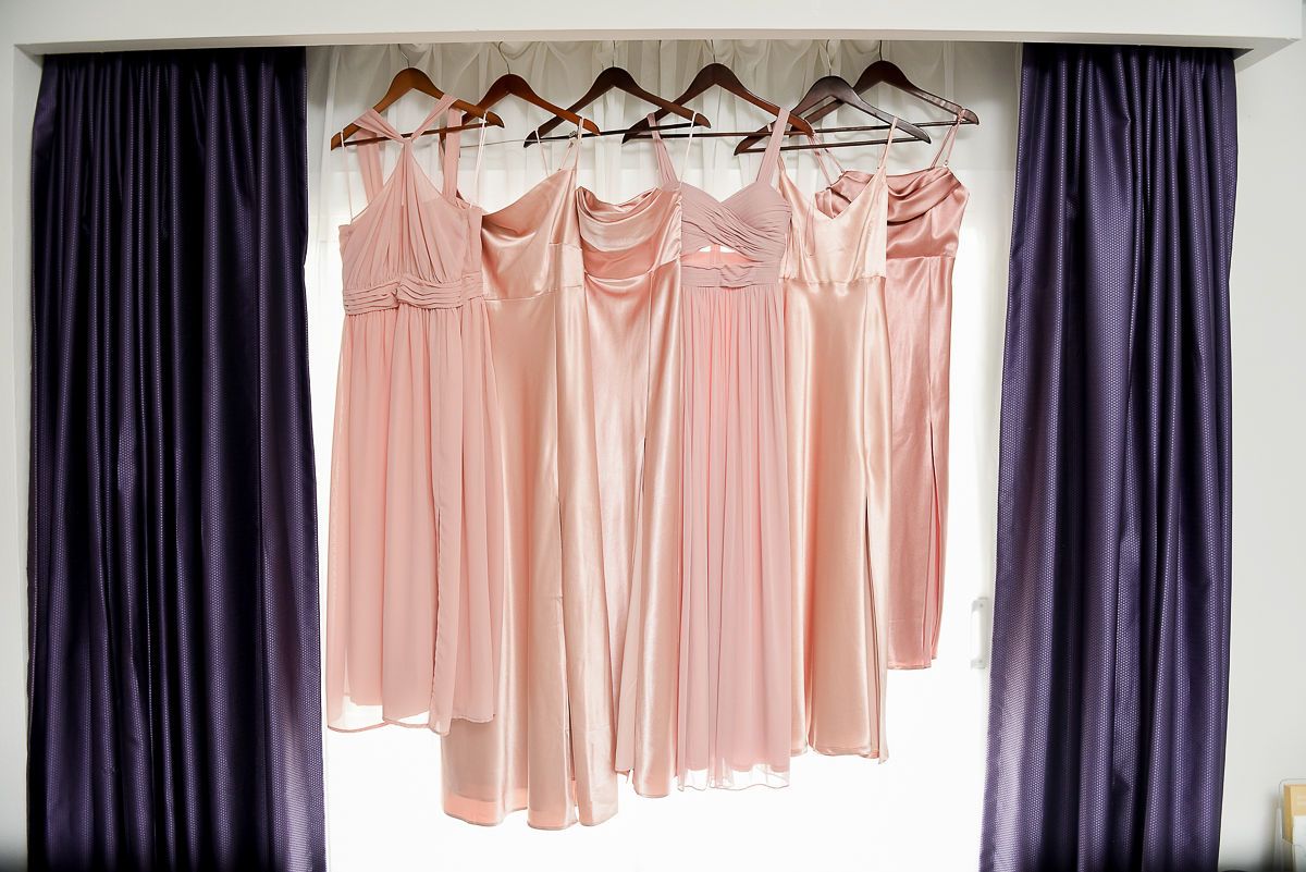 Five blush pink bridesmaid dresses hanging on wooden hangers between purple curtains.