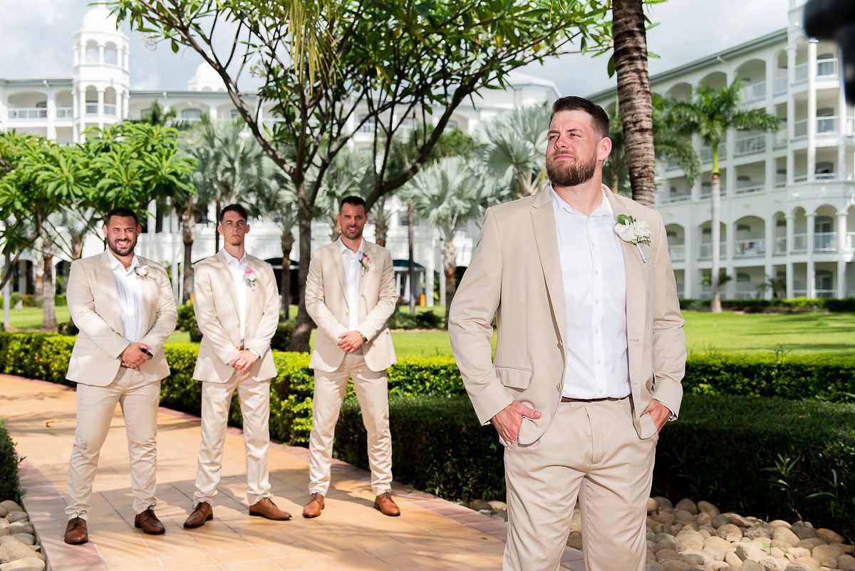 Groom in tan suit stands confidently outdoors at resort with groomsmen in matching suits in background.