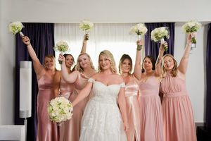 Bride in white lace gown poses with six bridesmaids in pink dresses holding white bouquets aloft in celebration.