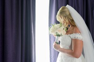 Blonde bride in lace off-shoulder gown holding white bouquet near draped curtains.