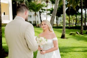 Bride in white off-shoulder gown holds bouquet facing groom in beige suit at tropical outdoor wedding venue.