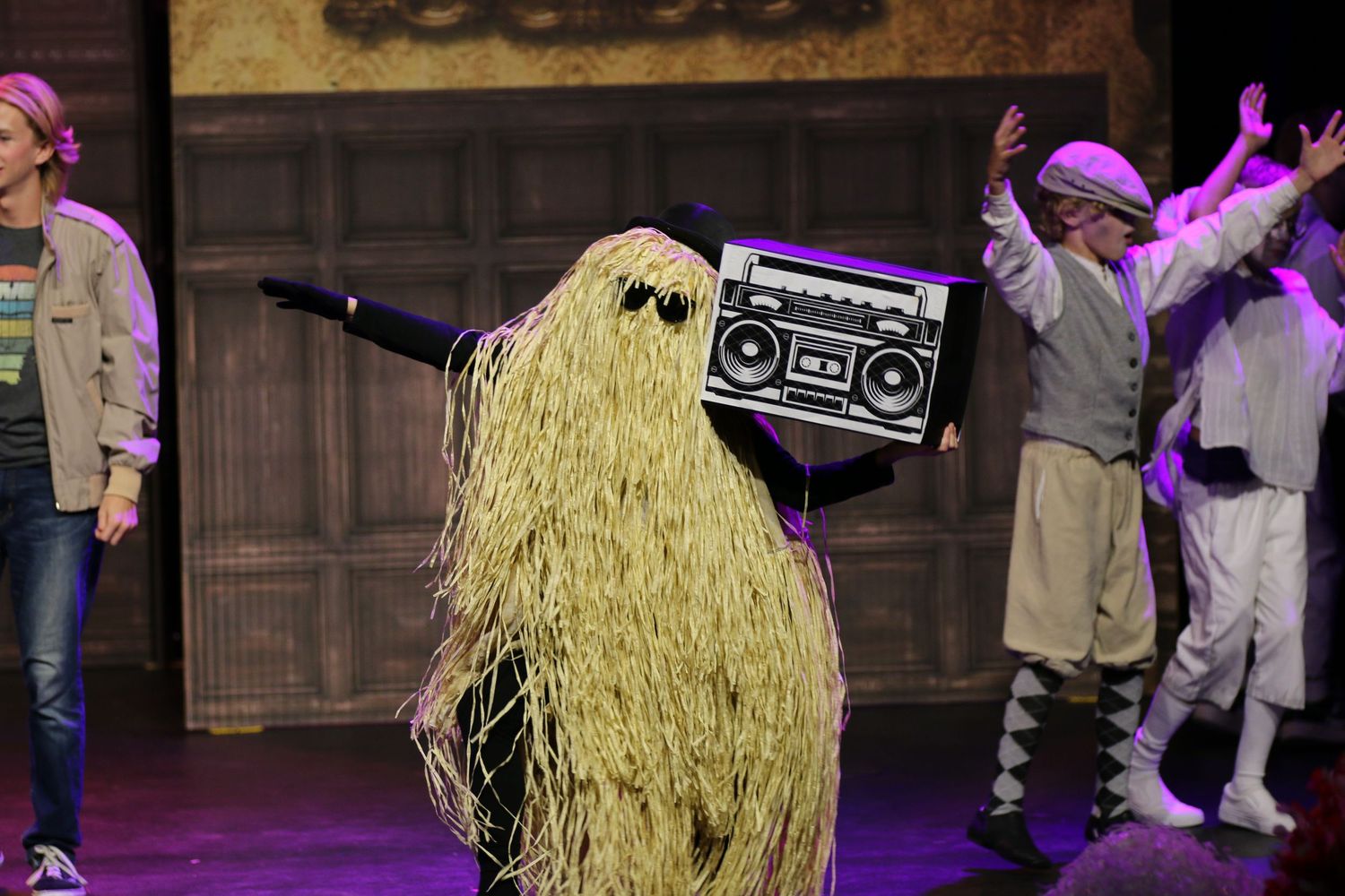 Performer in a shaggy costume carries a boombox on stage during a colorful theatrical production.