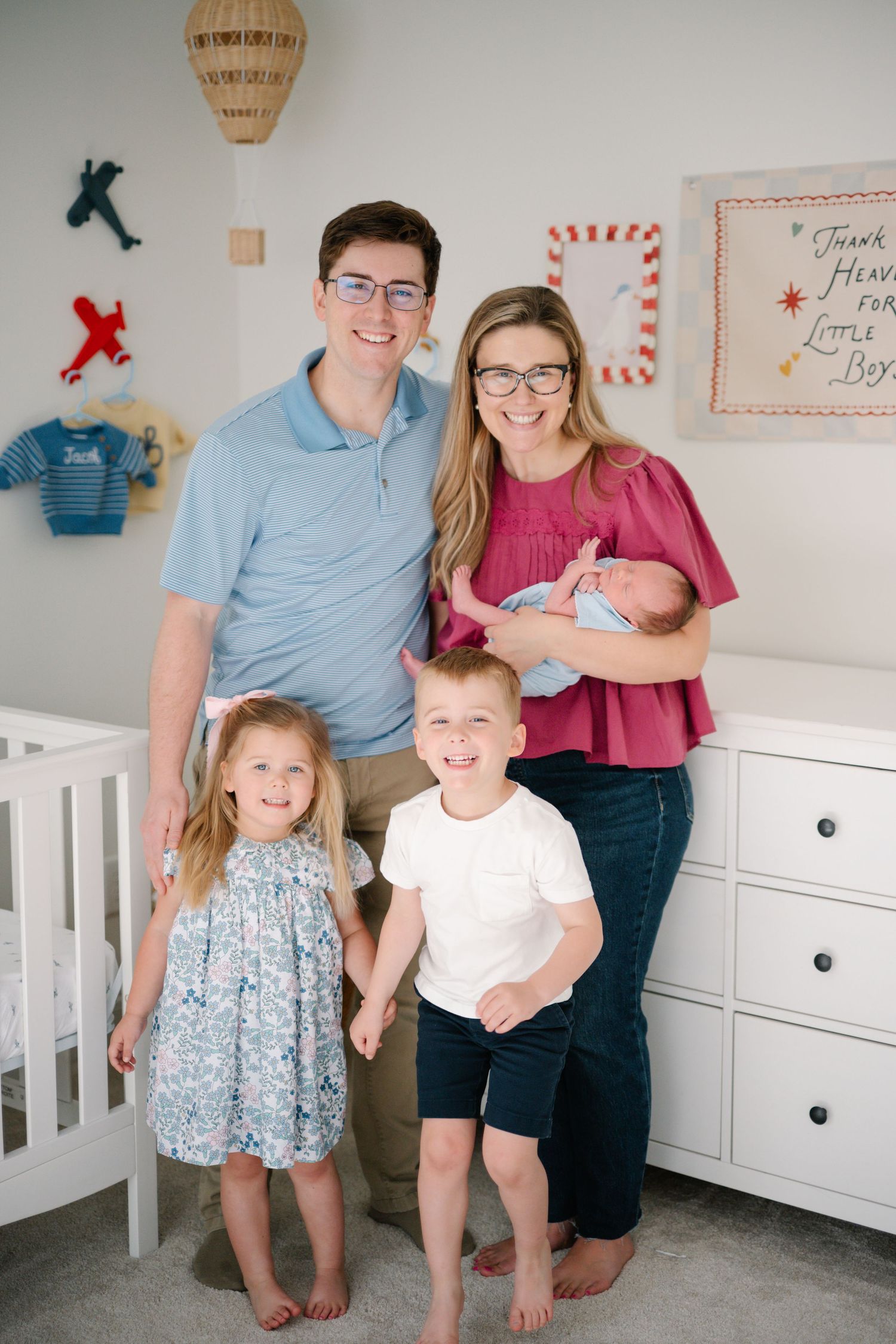 Family of five posing in a baby boy nursery with airplane decor, parents holding newborn with two young children.