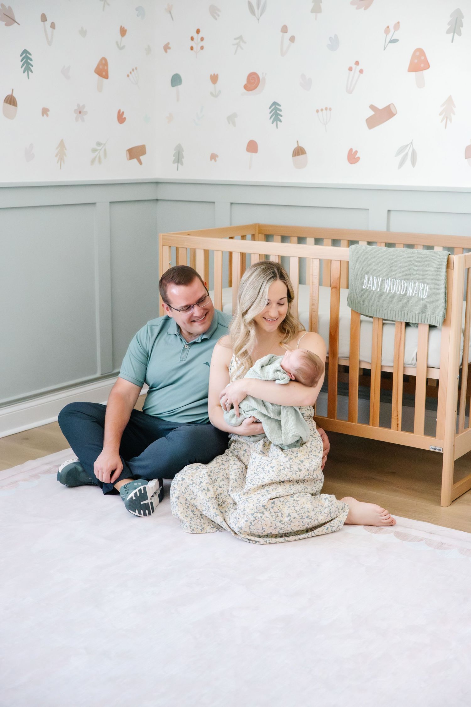 Parents sitting on nursery floor beside wooden crib, mother cradling newborn in floral blanket, father smiling warmly.