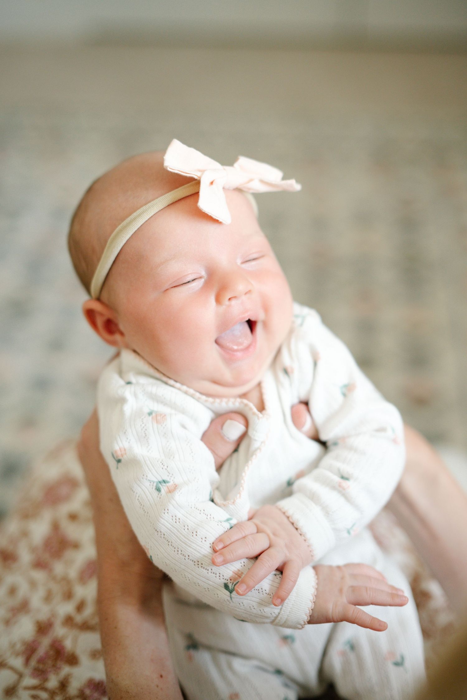 A joyful newborn baby girl wearing a white floral onesie and pink bow headband laughs while being held.