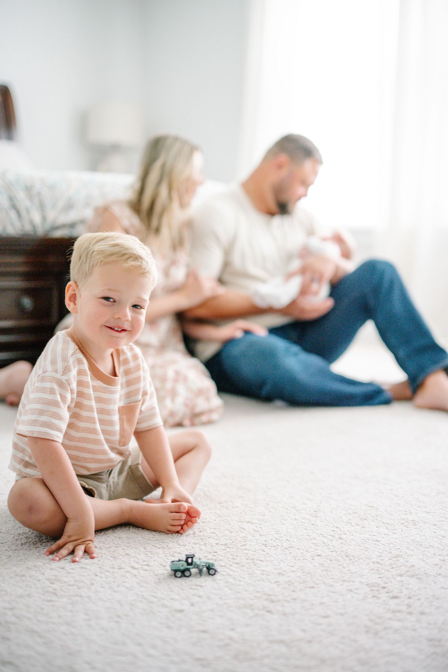 Young boy sits cross-legged on carpet with toy truck while parents hold newborn baby in bright bedroom.