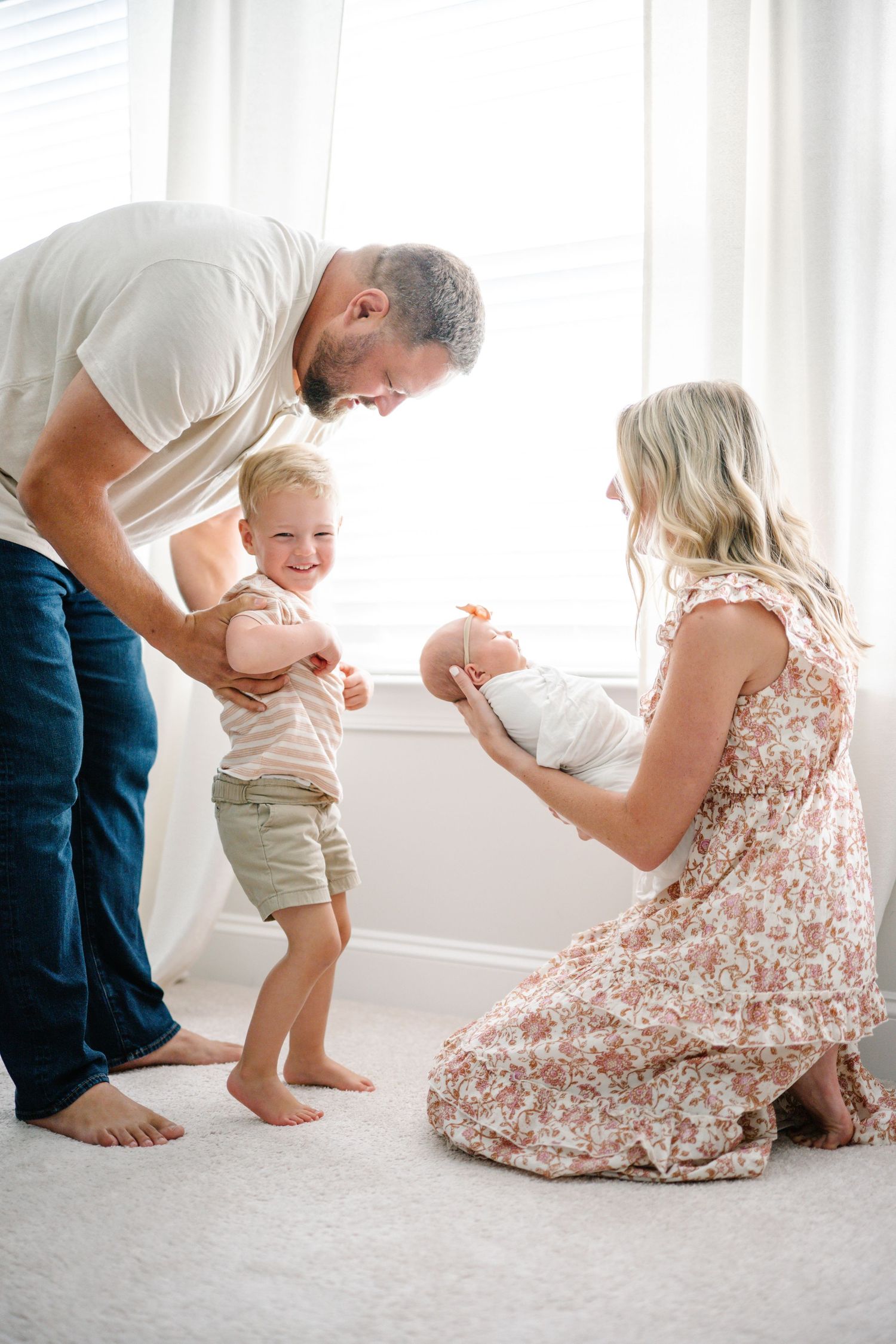 A happy family with a toddler boy meeting their newborn baby in a bright, airy room.