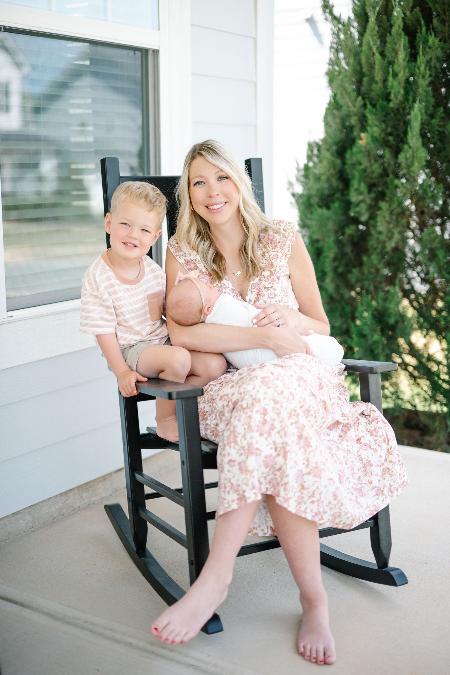 A smiling mother in a floral dress sits in a black rocking chair holding a newborn, with her young blond son beside her.