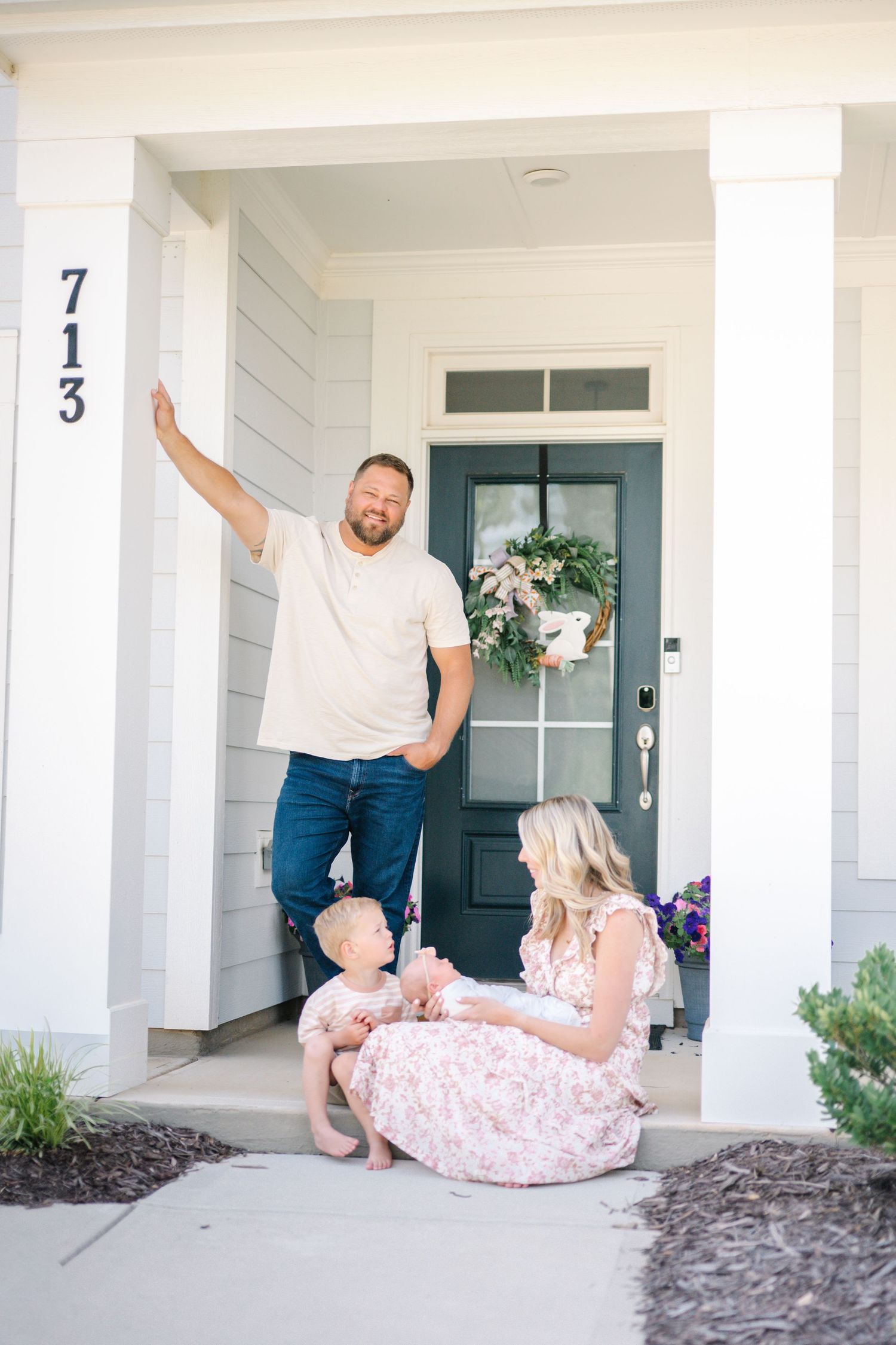 Family of three posing on front porch of home numbered 713, with dad standing, mom sitting, and toddler between them.