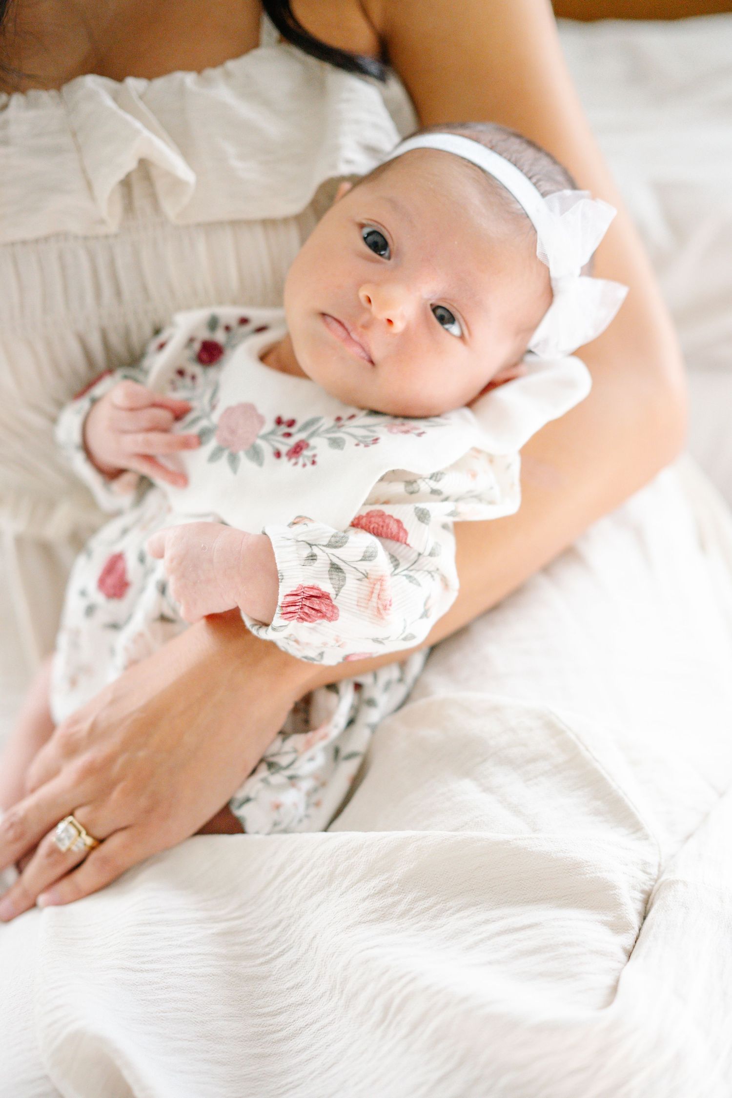 Newborn baby girl in floral outfit and white bow headband cradled in mother's arms on white bed.