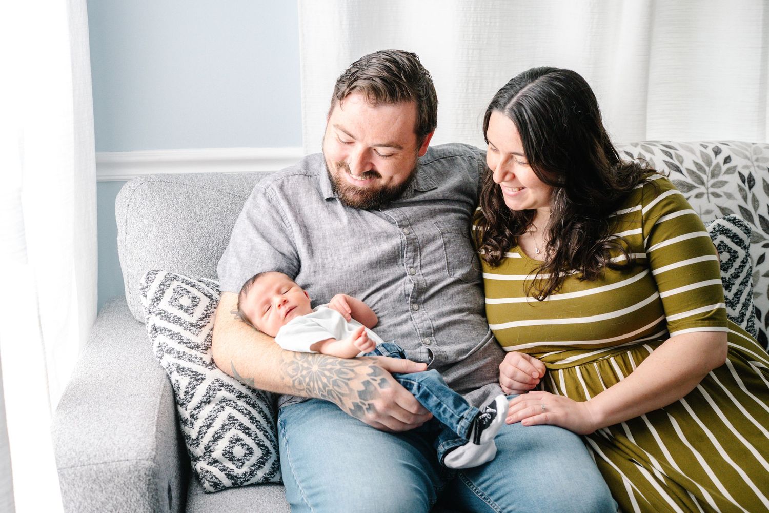 A couple sits on a gray sofa smiling while the man holds their newborn baby wrapped in a white blanket.