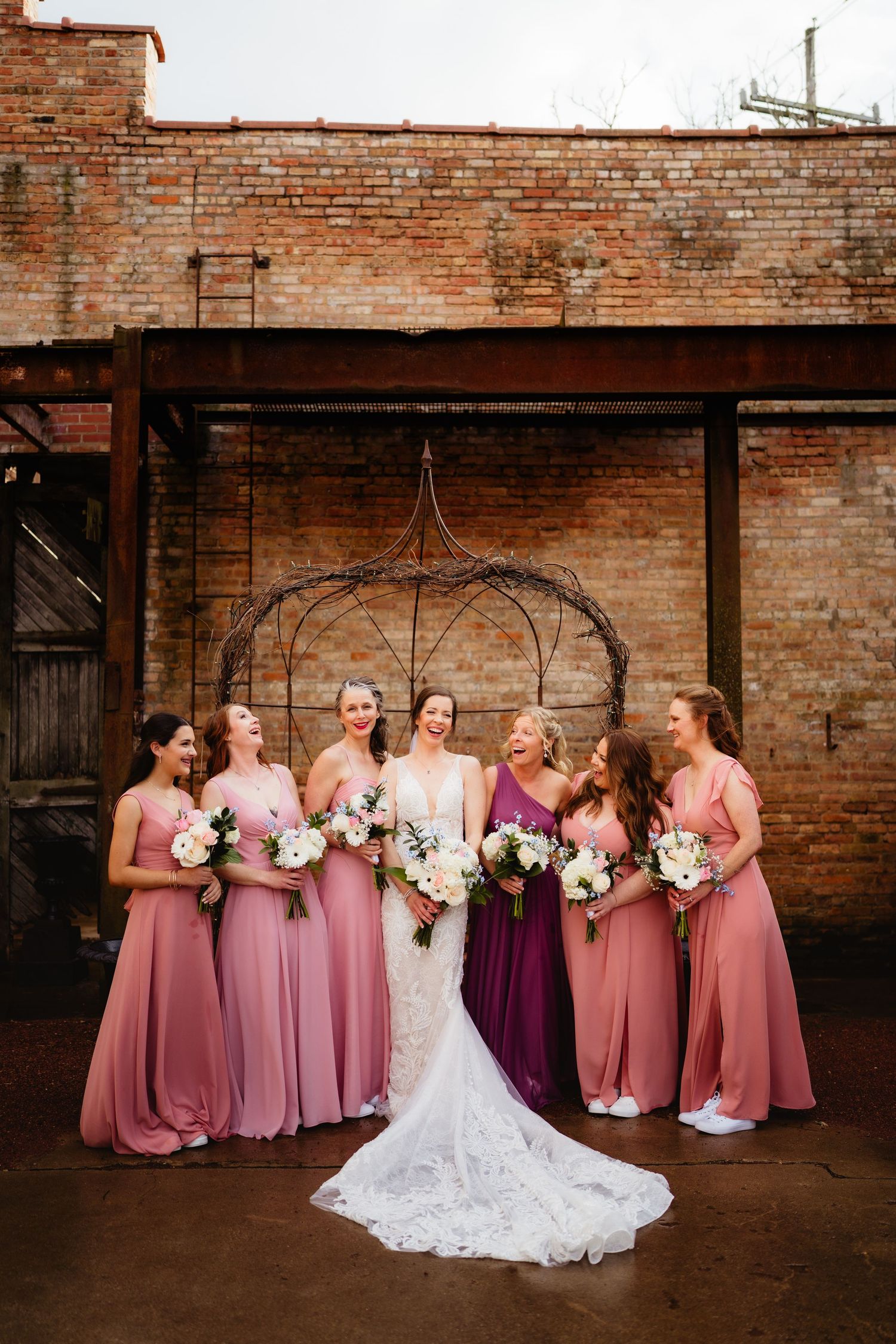 Bride in white lace gown with bridesmaids in pink and purple dresses holding bouquets before brick wall backdrop.