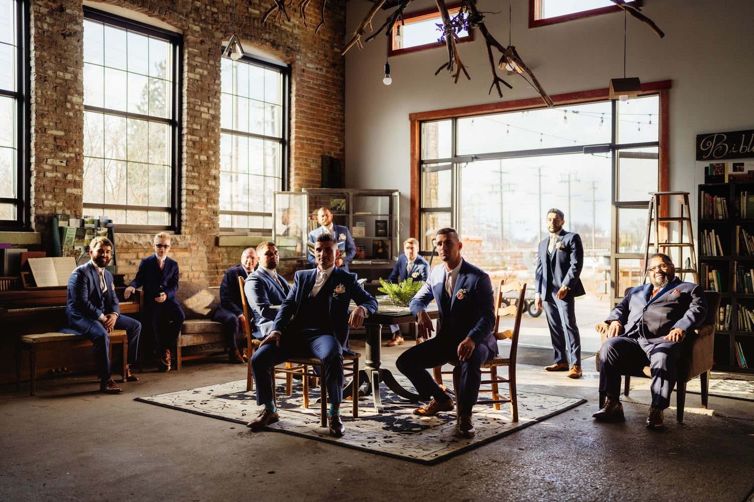 Groomsmen in navy suits gathered in rustic industrial venue with exposed brick walls and large windows.