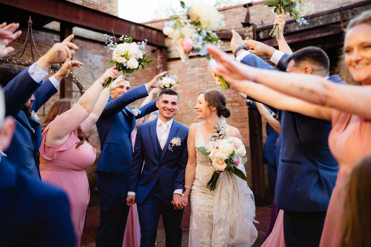 Bride and groom walk through cheering wedding party in pink and navy attire, holding bouquets outdoors.