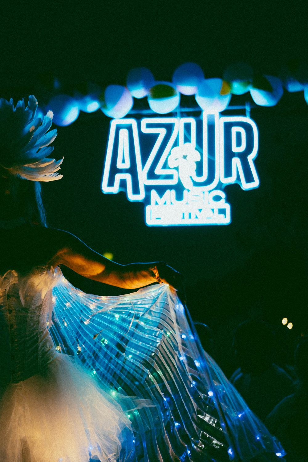 Performer in illuminated costume at Azur Music Festival with glowing neon sign backdrop.