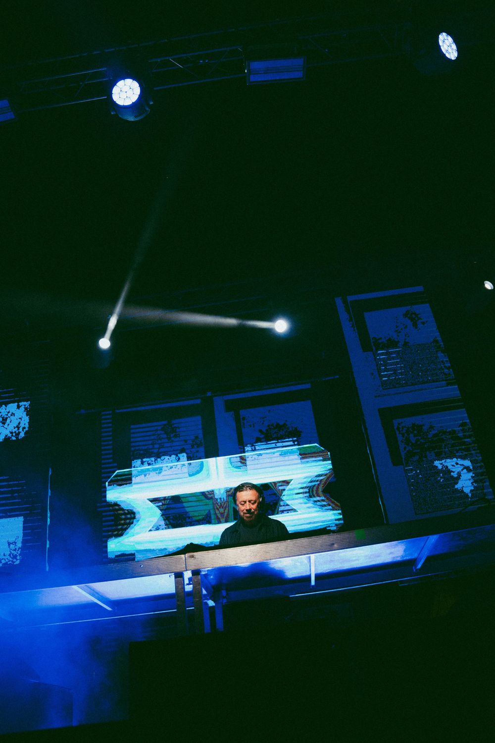 DJ se produisant sur scène sous des projecteurs bleus et blancs avec des écrans lumineux dans une salle de concert sombre.