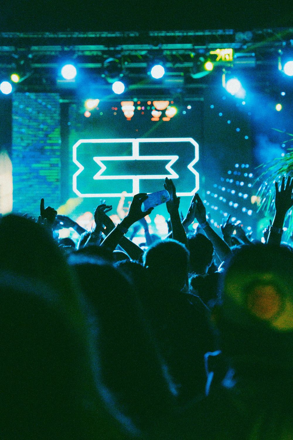Crowd cheering at a live concert with green neon lights and a glowing logo on stage.