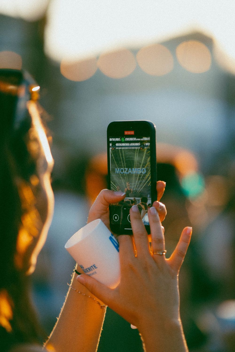 Femme tenant un smartphone lors d'un festival de musique, capturant une performance à l'écran à la lumière de l'heure dorée.