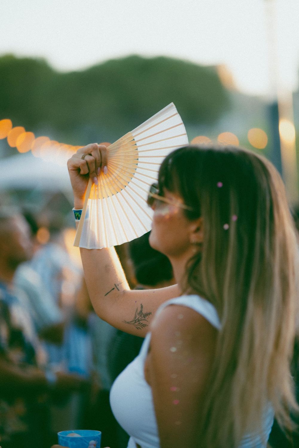 Femme tenant un éventail à main blanche lors d'un festival en plein air, portant des lunettes de soleil et montrant des tatouages