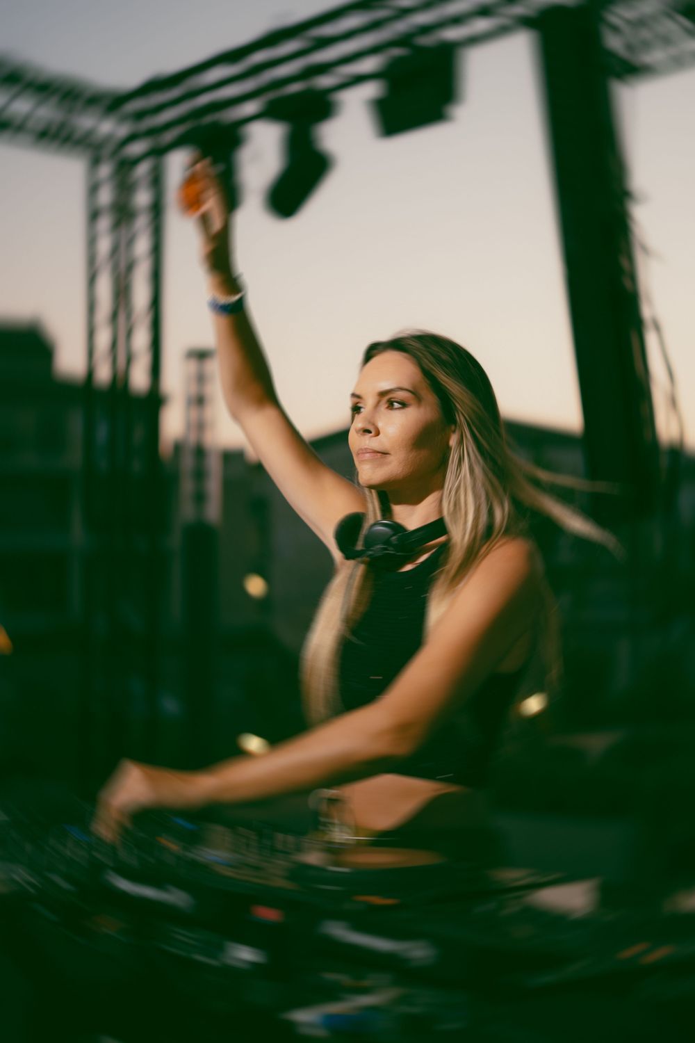 Female DJ with headphones around neck raises hand while performing at outdoor music festival at dusk.