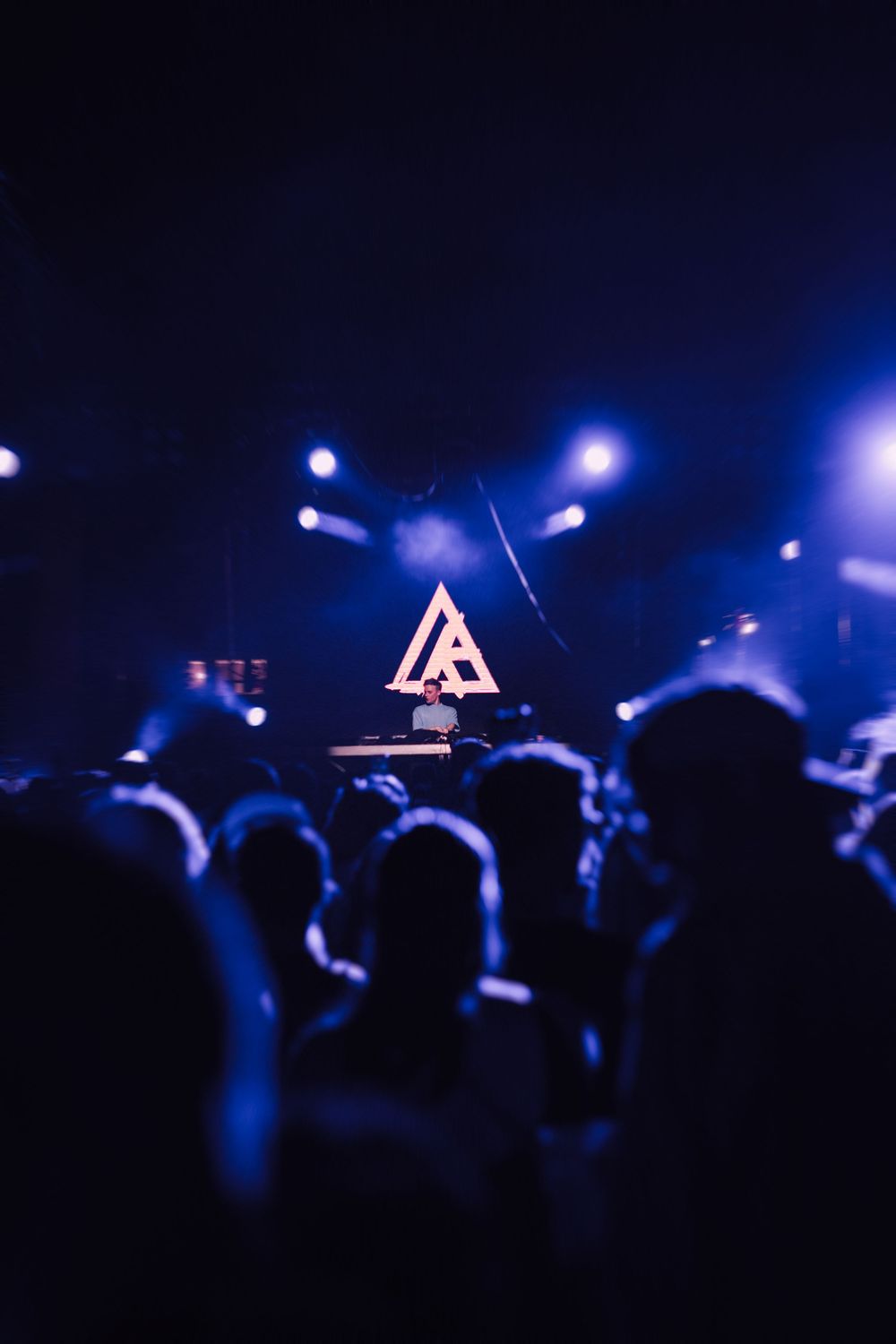A DJ performs on stage with a glowing triangle logo behind them, surrounded by a crowd in blue-lit concert venue.