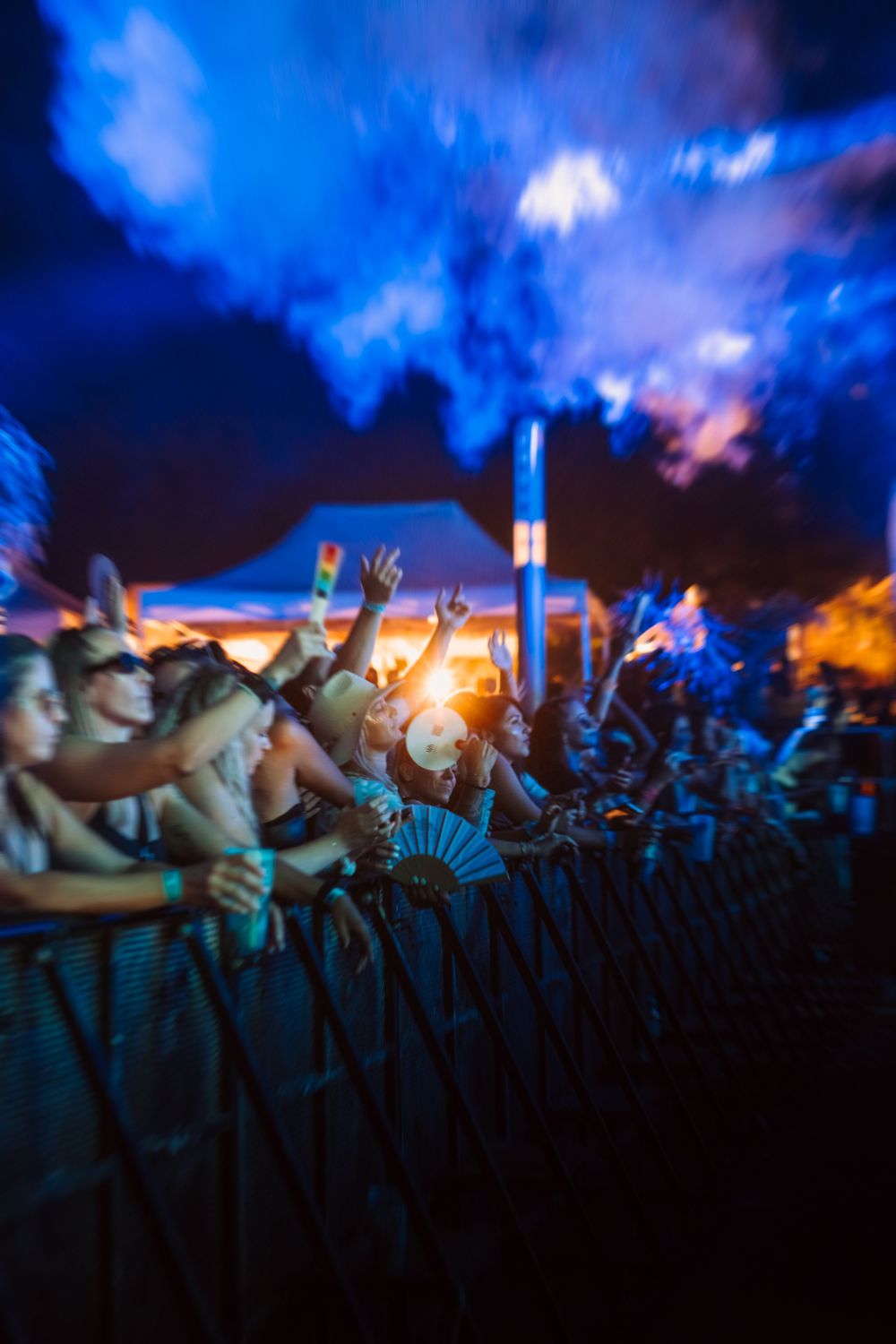Excited concert crowd behind metal barriers under blue and orange lights at an outdoor music festival at night.