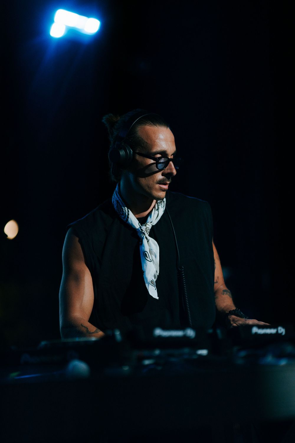DJ performing at night with Pioneer DJ equipment, wearing headphones and black eye mask under dramatic stage lighting.