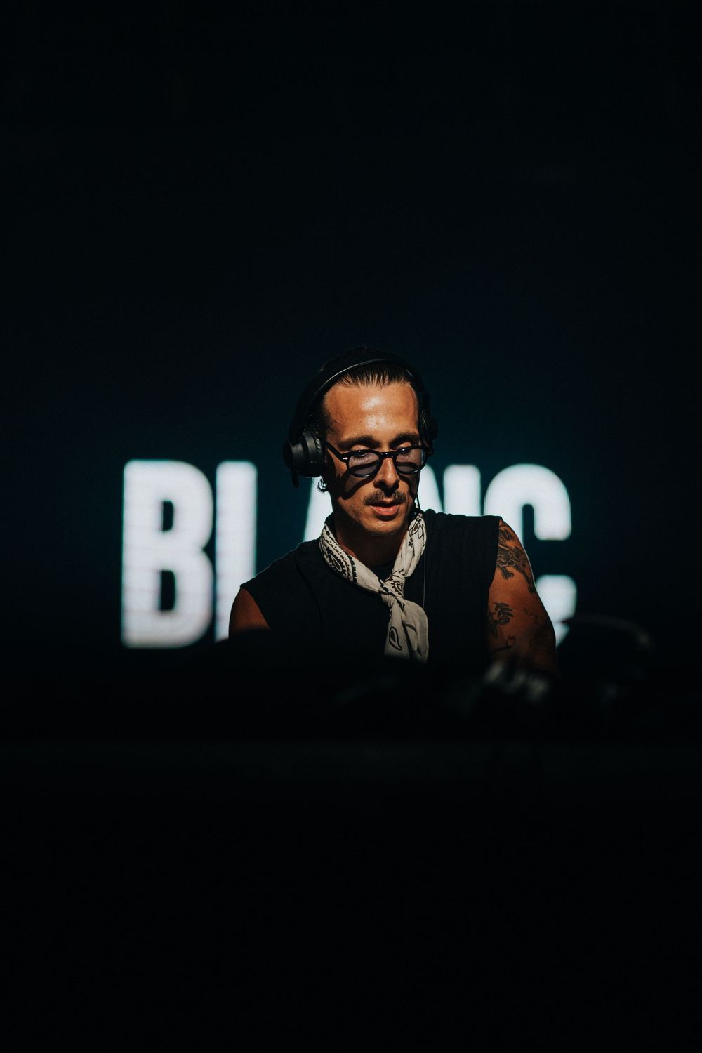 DJ performing at Blanc event, wearing glasses and headphones, with tattoos visible in dramatic dark lighting.