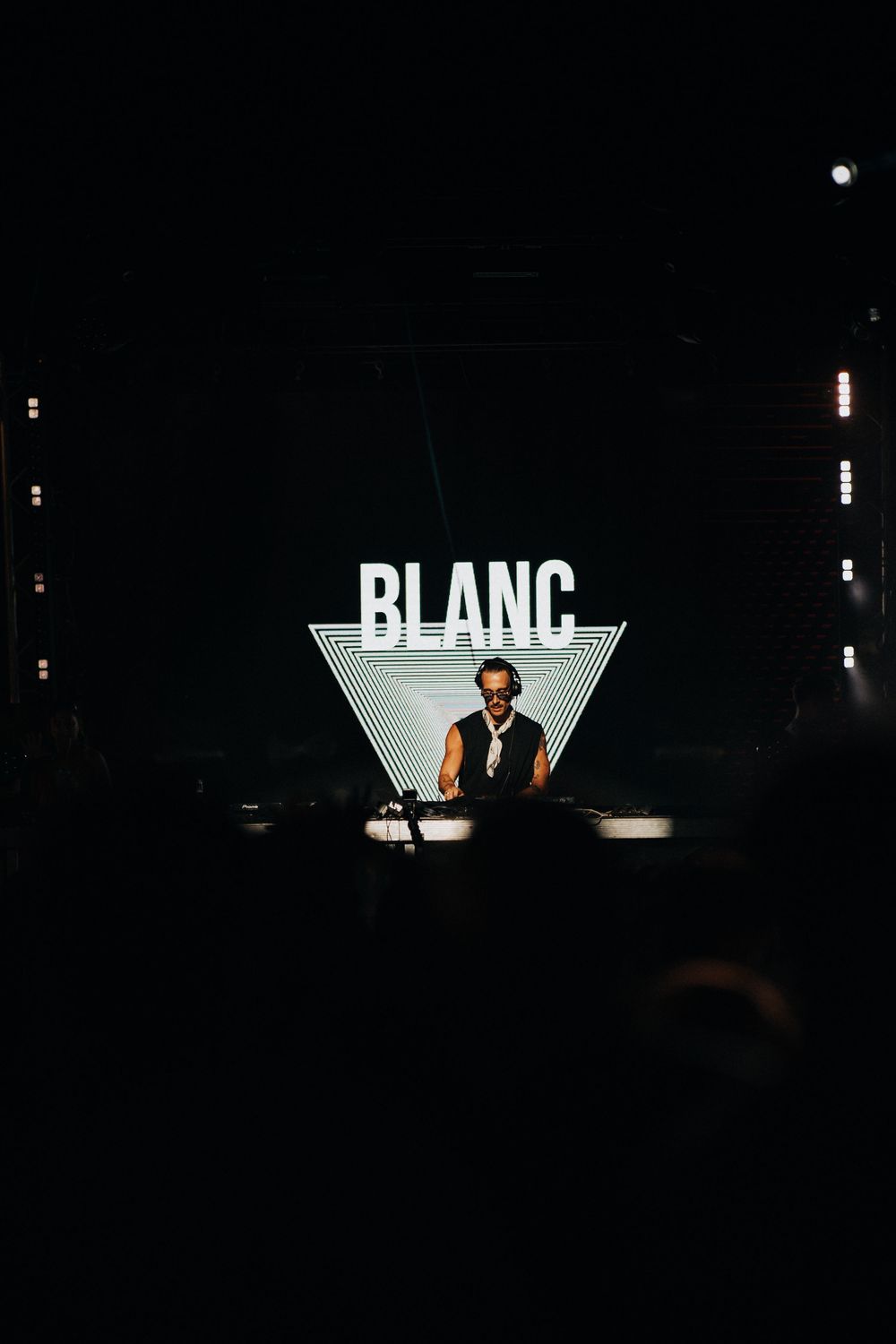 DJ performing at Blanc event under glowing triangle logo sign in dark concert venue.