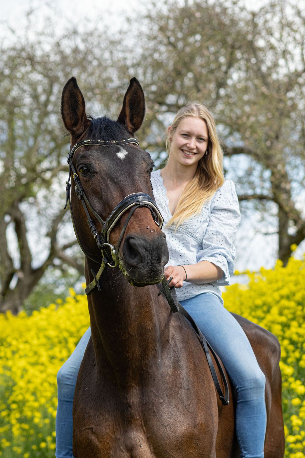 Glimlachende vrouw rijdt op een donkerbruin paard langs een veld met gele bloemen.