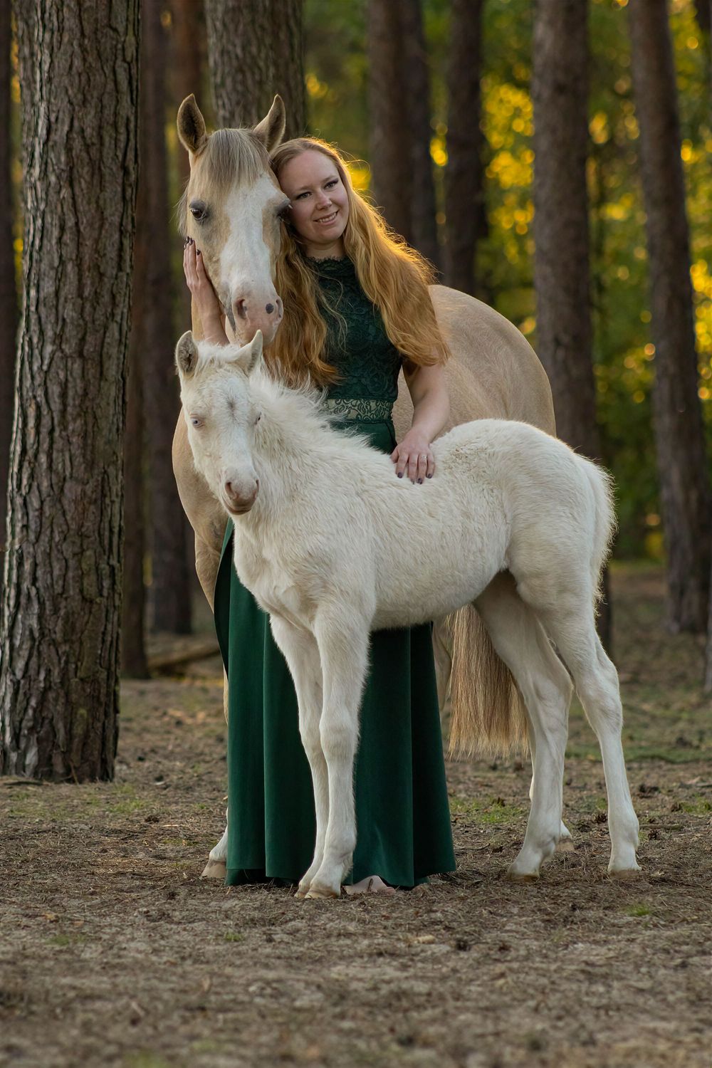 Vrouw in een lange groene jurk omhelst een wit veulen in een bos.