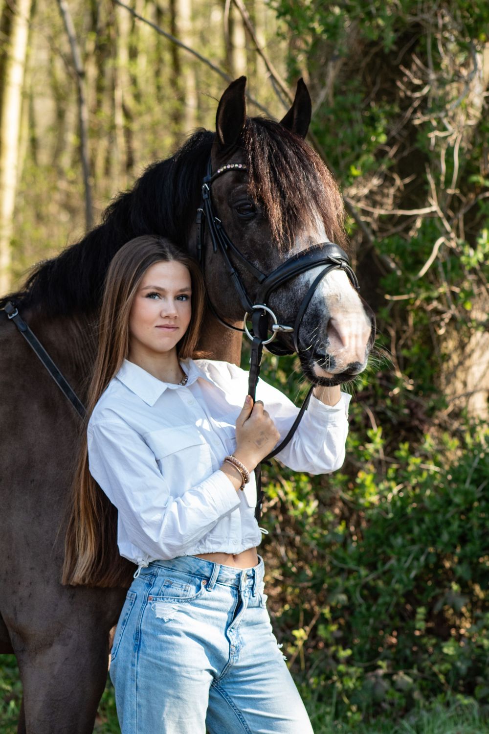 Jonge dame in een witte blouse staat naast een donkerbruin paard met een witte bles in een bos.