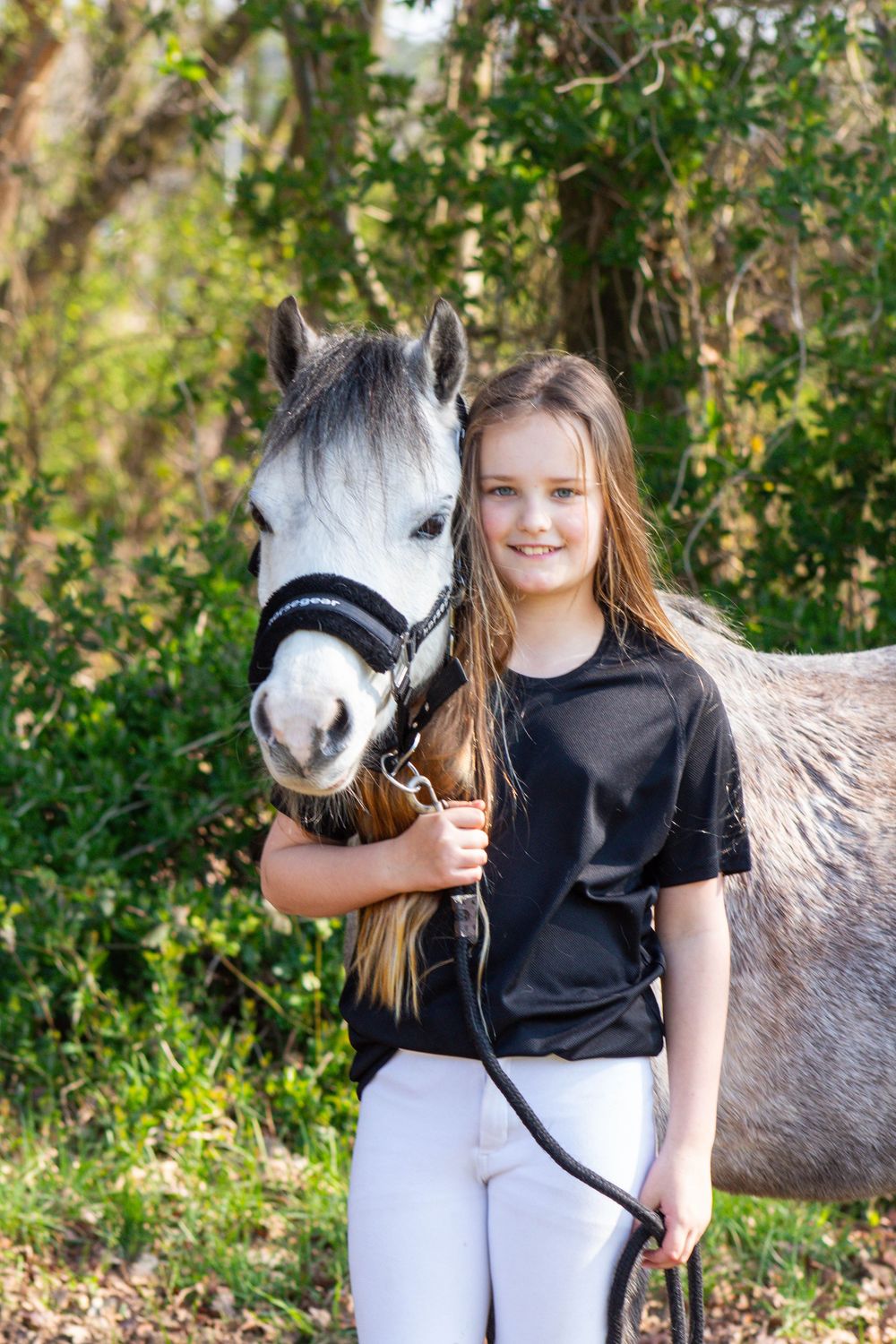 Glimlachende jonge vrouw staat naast een grijs paard met een halster, voor een achtergrond van groen struikgewas.