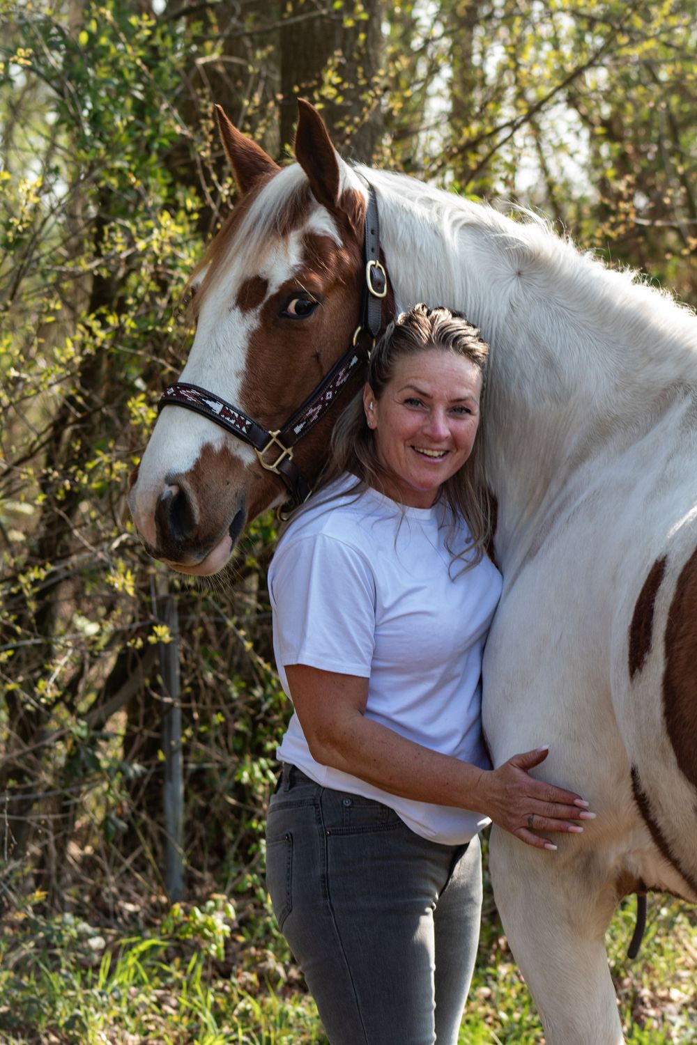 Mevrouw met blond haar, houdt haar bonte paard op de borst vast en kijkt naar de camera. Bossen op de achtergond