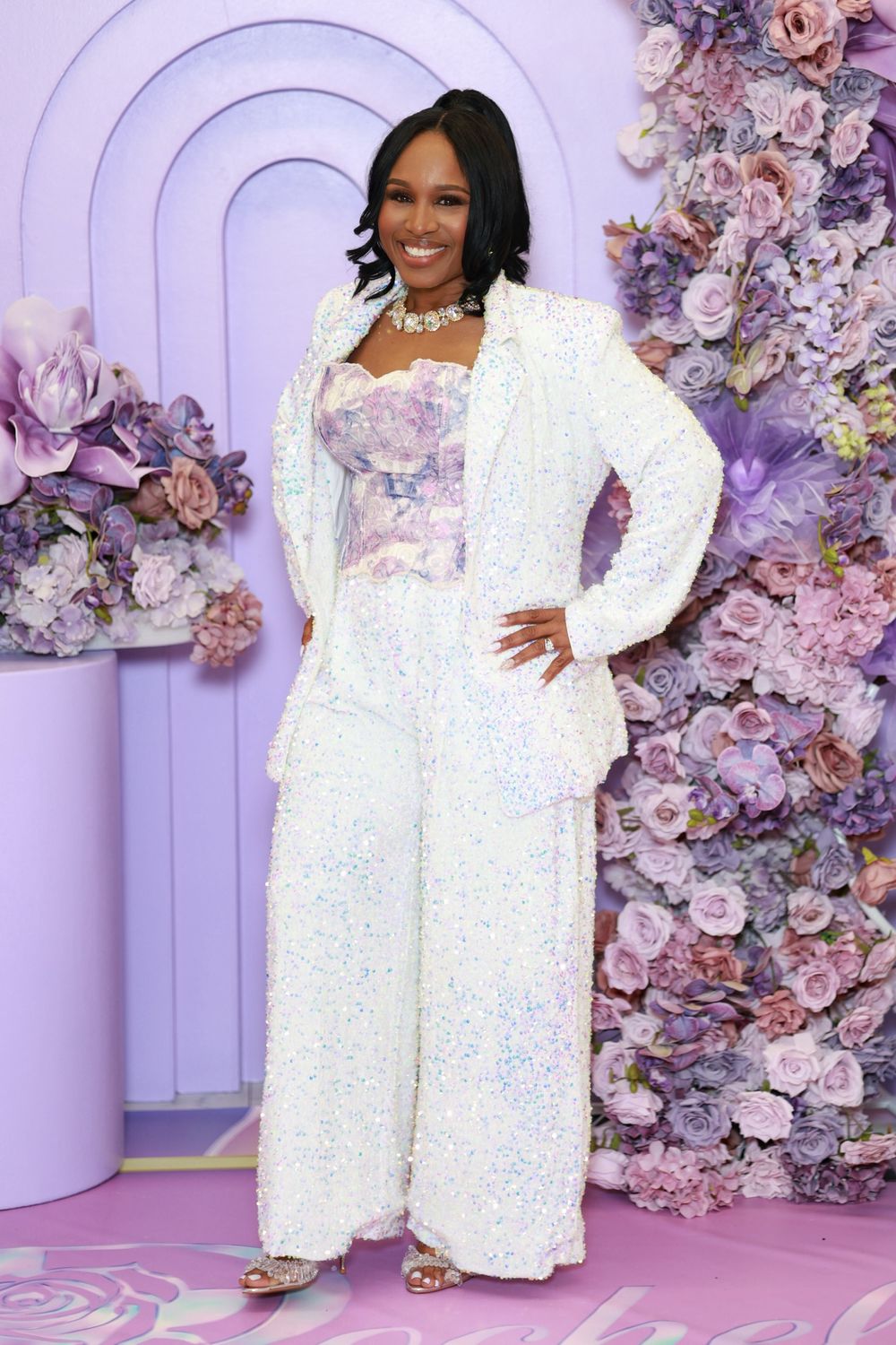 Woman in white sequin blazer and wide-leg pants poses before floral wall backdrop at elegant event.