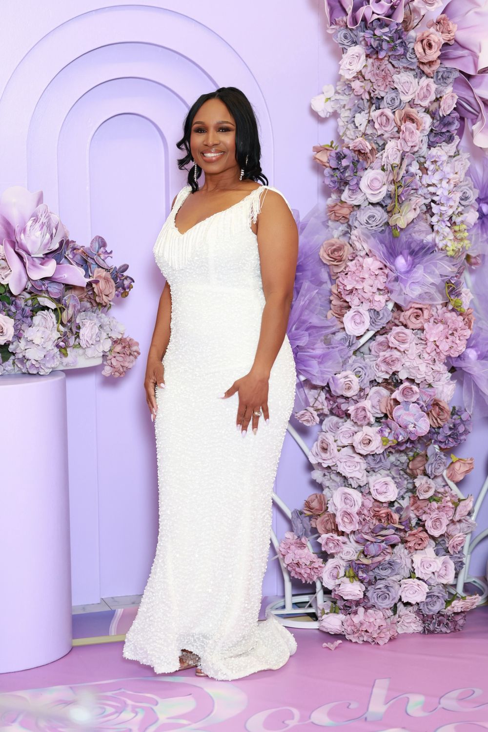 Woman in white beaded gown posing before purple floral backdrop at elegant event.
