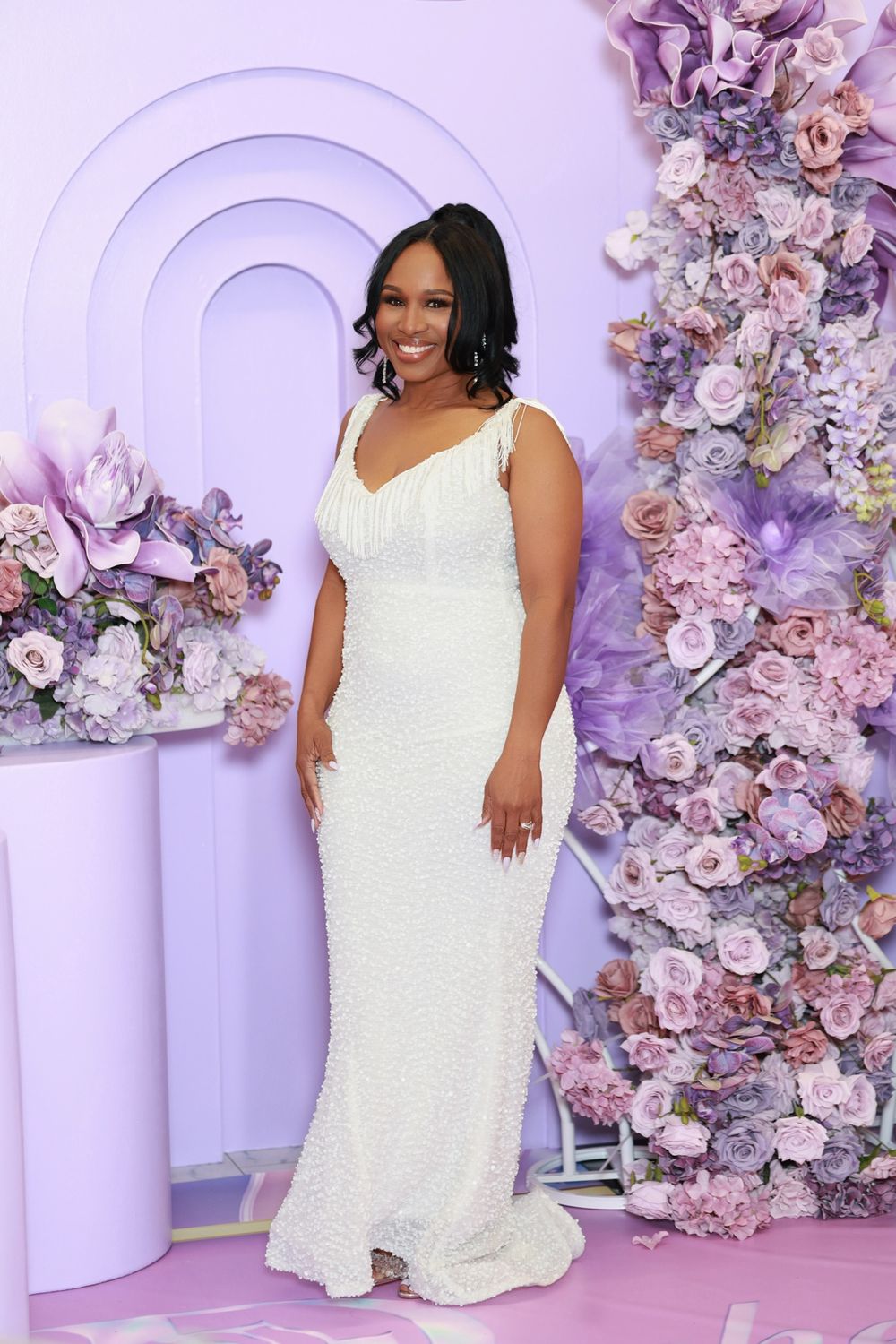 Woman in white sequin gown poses before purple floral backdrop at elegant event.