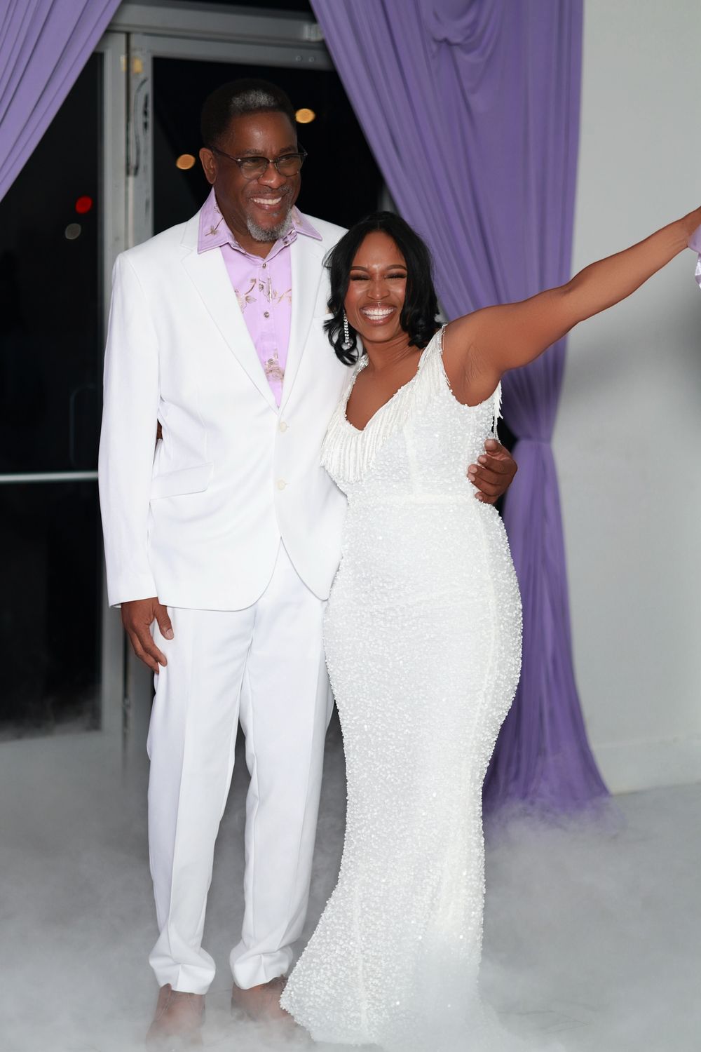 Couple in white formal attire posing elegantly at an event with purple drapes and atmospheric fog.