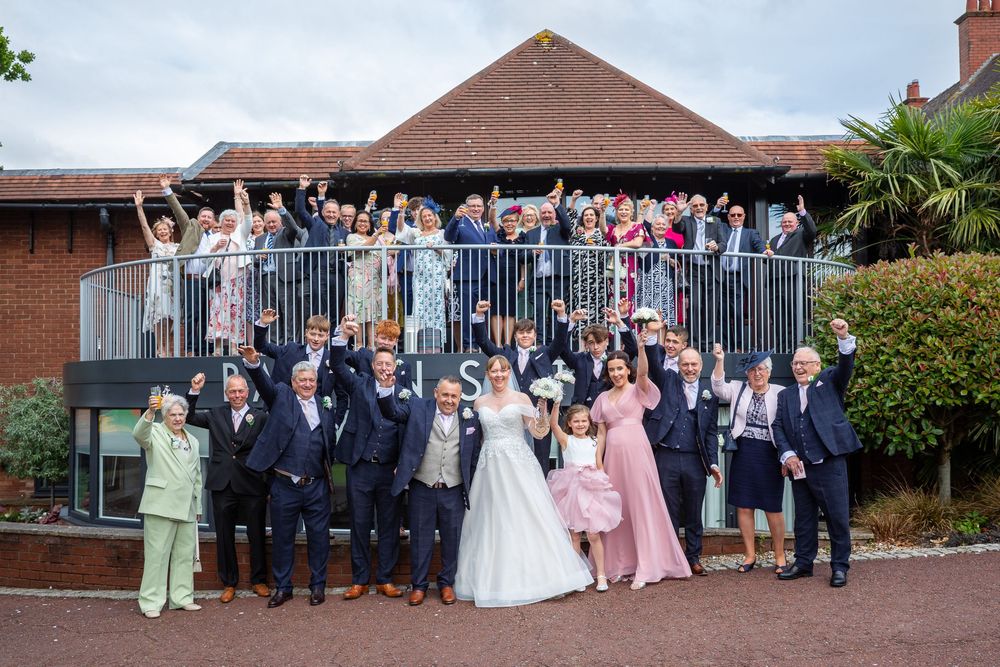 Newlyweds and wedding guests celebrate together outside a brick wedding venue raising hands.