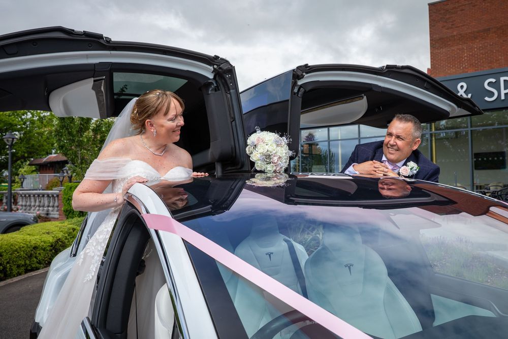 Bride and groom share a moment beside a ribbon-decorated luxury wedding car outside a spa.