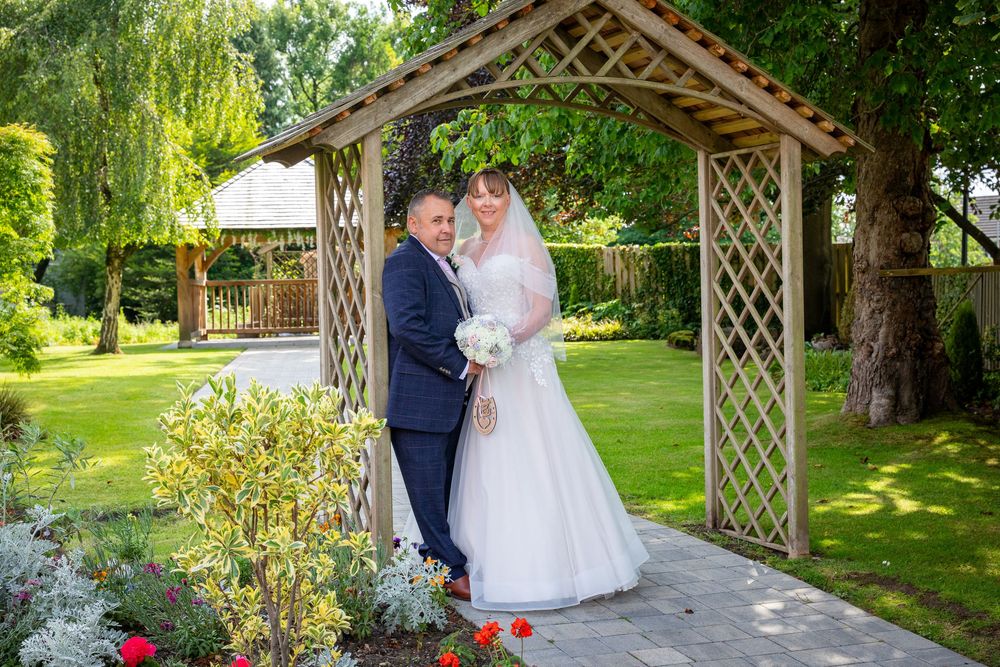 Newlyweds pose romantically under a wooden garden arbor surrounded by lush greenery.