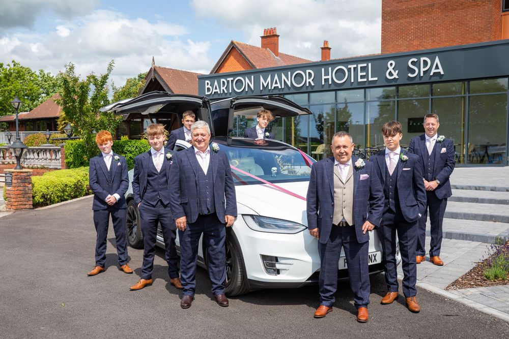 Groomsmen in navy suits pose with a white Tesla outside Barton Manor Hotel and Spa.