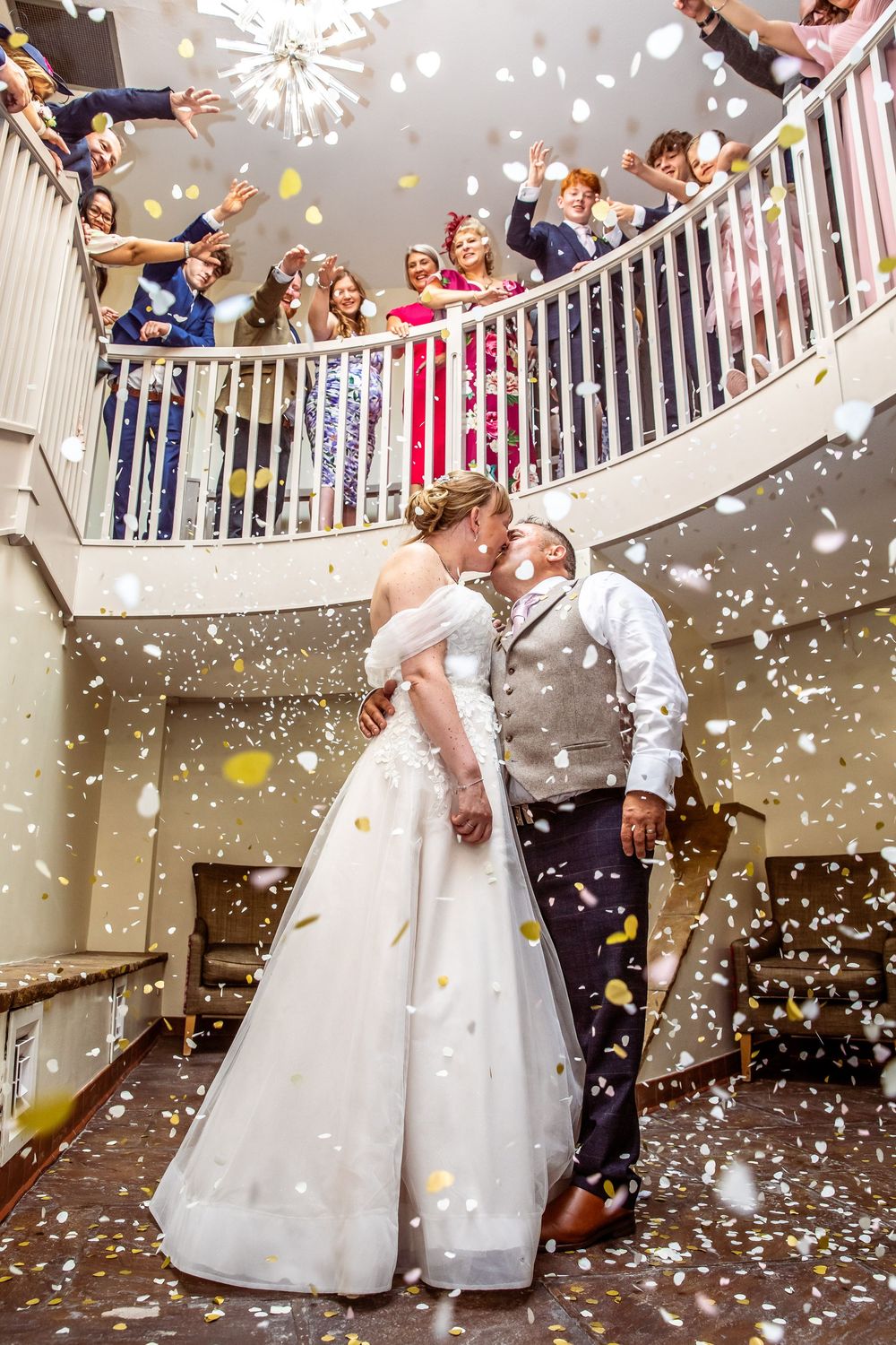 Newlyweds share a kiss under a staircase as confetti falls and guests cheer from balcony.